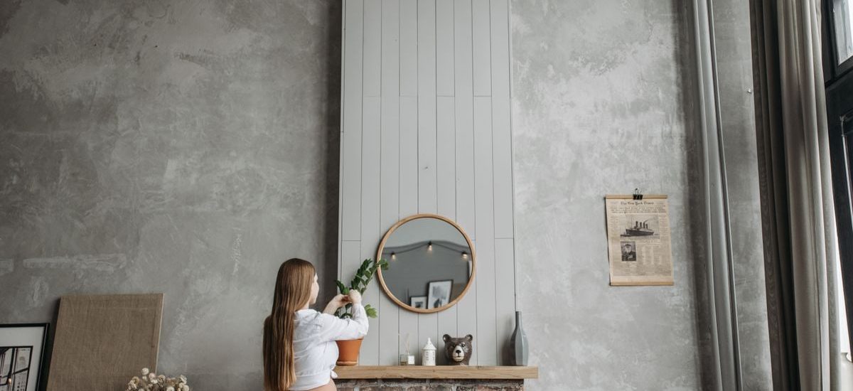 Pregnant woman in cozy living room arranging potted plant on fireplace mantel with rustic decor.