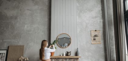 Pregnant woman in cozy living room arranging potted plant on fireplace mantel with rustic decor.