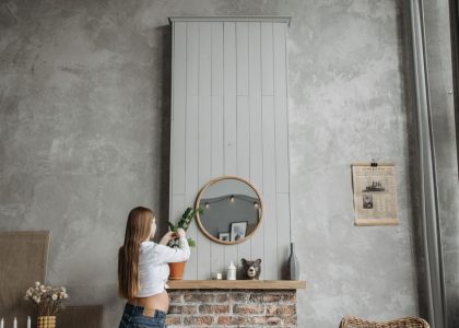 Pregnant woman in cozy living room arranging potted plant on fireplace mantel with rustic decor.