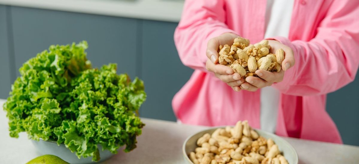 Hands presenting peanuts with a bowl, lettuce, avocado, and tomato nearby on kitchen counter.