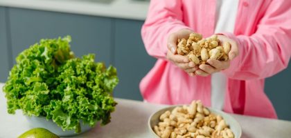 Hands presenting peanuts with a bowl, lettuce, avocado, and tomato nearby on kitchen counter.